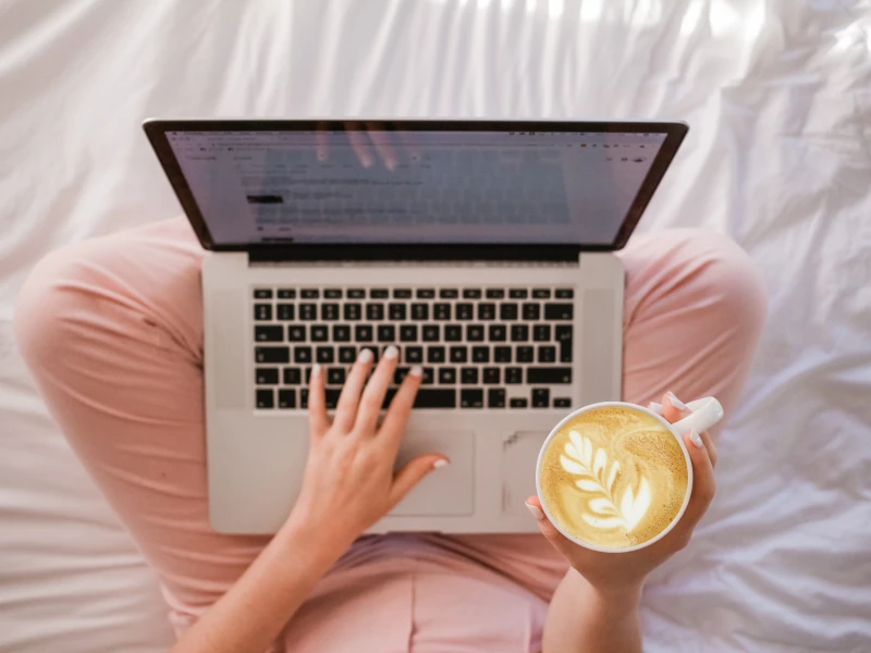 ベッドの上であぐらをかいた上にノートパソコン乗せ、左手はタイピングし右手はコーヒーを持っている様子を真上のふかん視点から写した写真。